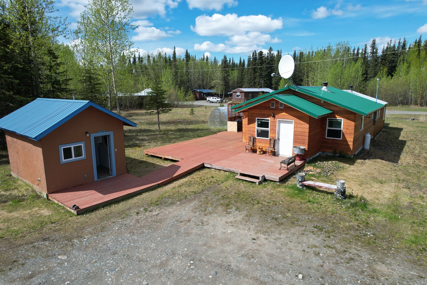 Aerial view of a rural property showing two wooden cabins connected by a red deck: a green-roofed main house with a satellite dish and a blue-roofed shed nearby, surrounded by trees and gravel yard.